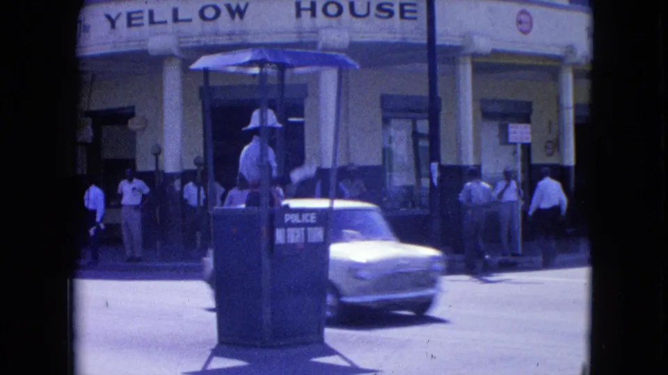 A white compact car moves past the traffic-control booth as the officer stands outside the Yellow House in 1960s Kingston.