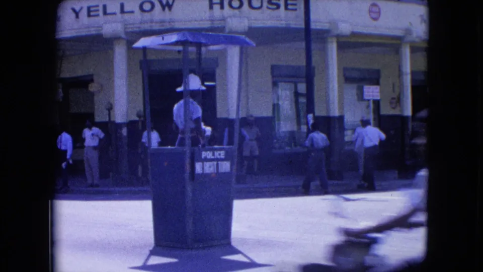 In Kingston, Jamaica, a white-uniformed police traffic guard stands in a no-right-turn booth outside Yellow House in 1964 street footage.