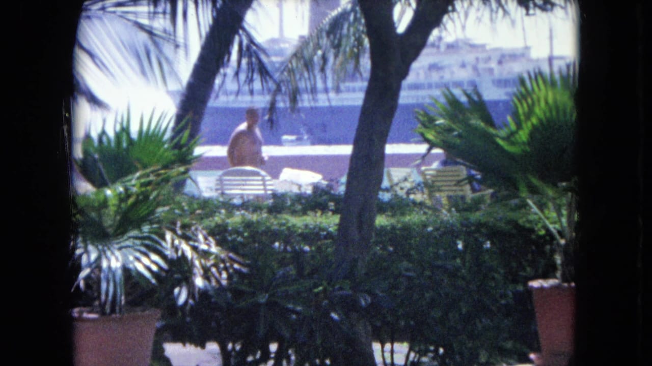 Man in swim trunks walks across a patio behind palms and lounge chairs.
