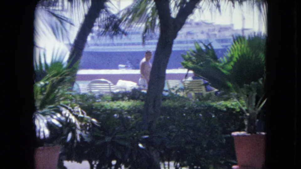 1964 Kingston, Jamaica 8mm view of a man moving through a shaded tropical patio with chairs and a cruise ship behind.