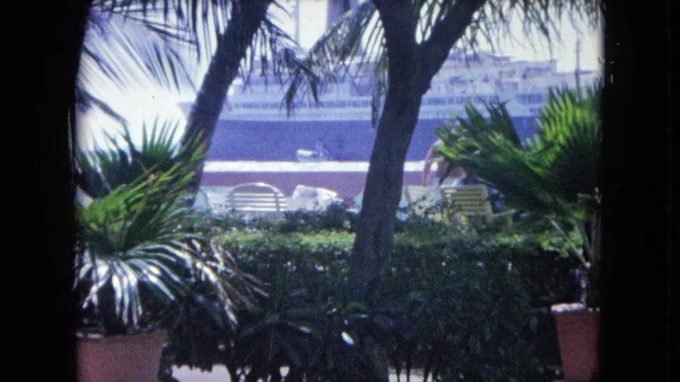 1964 Kingston, Jamaica frame looking through palms toward patio furniture, dense greenery, a person, and the ship in the background.