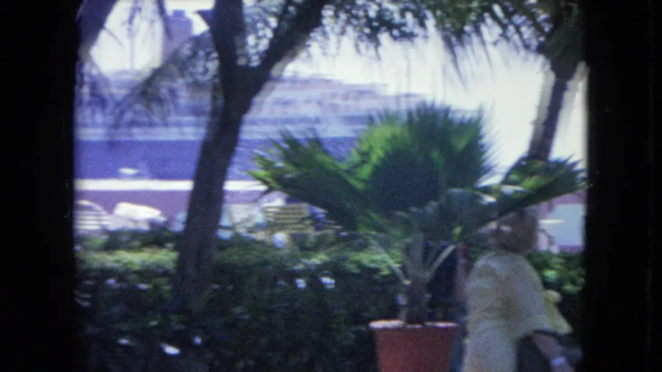 1964 Kingston, Jamaica coastal garden scene with a woman passing at frame edge, palm foliage, patio furniture, and a harbor ship beyond.