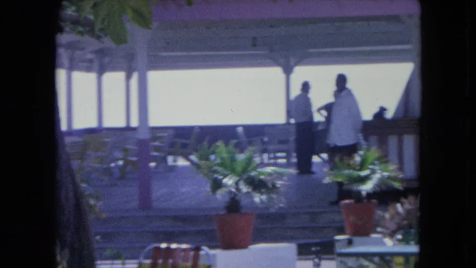 1964 Kingston home-movie frame showing two adults beneath a pavilion, with patio chairs, potted plants, columns, and an overexposed sky.