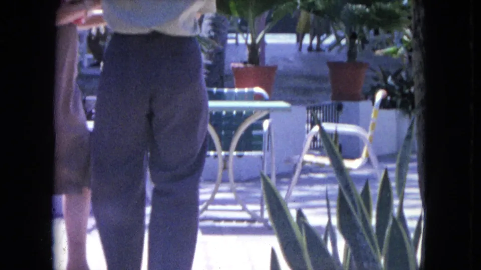 1964 Kingston, Jamaica patio frame showing a couple partly cropped while dancing among white chairs, tables, and potted tropical plants.