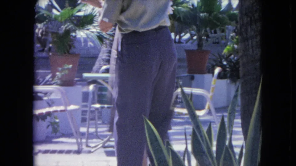 A 1964 Kingston, Jamaica home-movie frame shows a dancing figure on a sunlit patio framed by palms, chairs, tables, and potted plants.