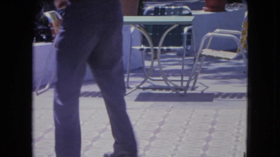 1964 Kingston home-movie frame with a man dancing across patterned patio tiles beside metal chairs and tables.