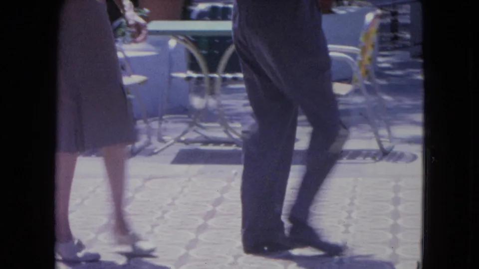1964 Kingston home-movie frame showing two adults mid-dance on a sunlit patio with metal furniture in view.