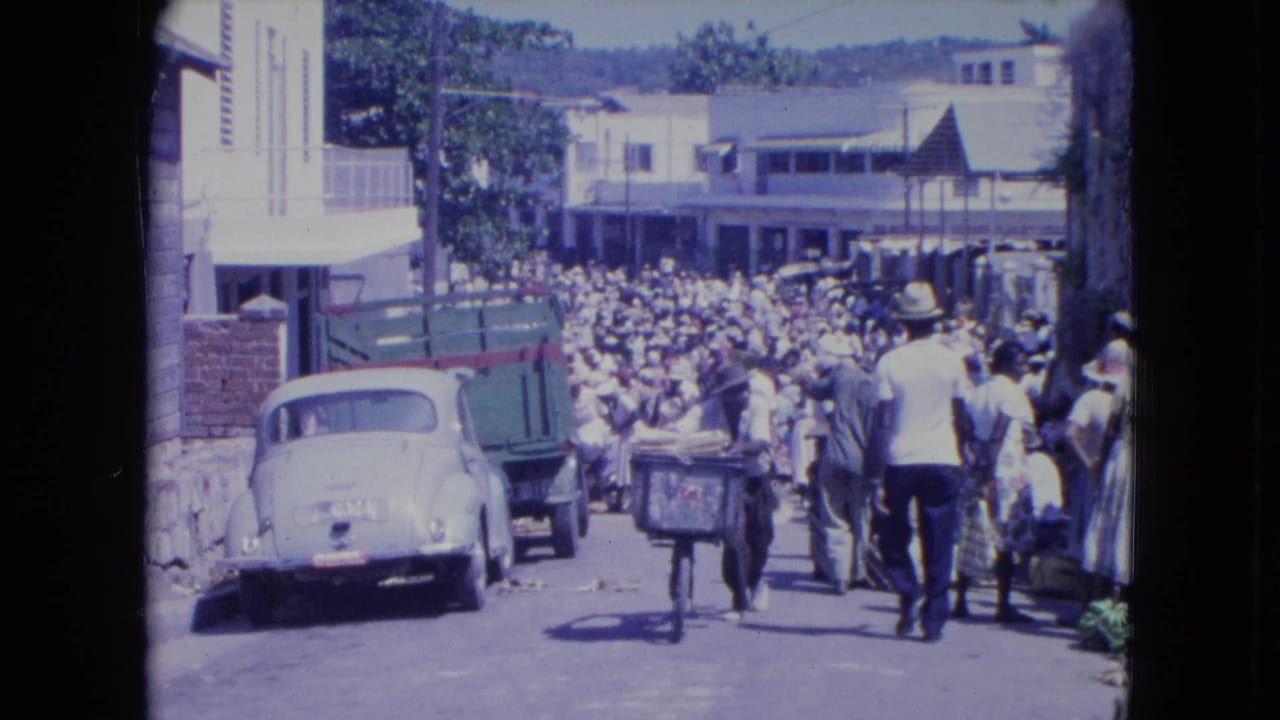 Crowded 1964 Kingston market street with people in hats, a parked Beetle, a green truck, and a cart moving through the road.