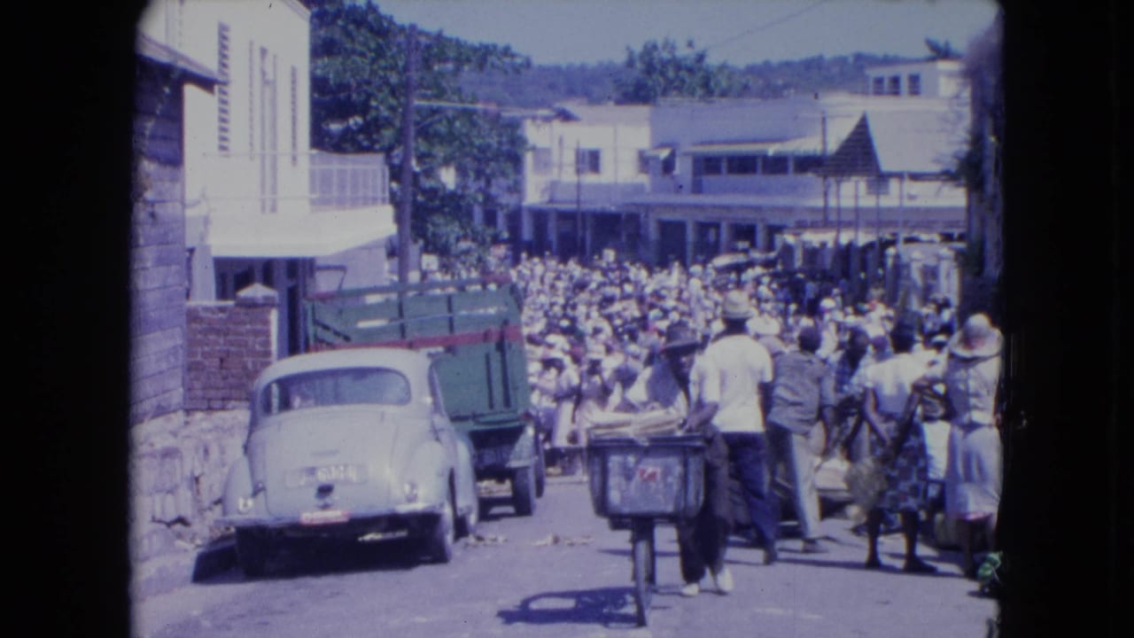 Elevated view of a crowded Kingston street market in 1964, with a Beetle, truck, carts, and pedestrians filling the road.