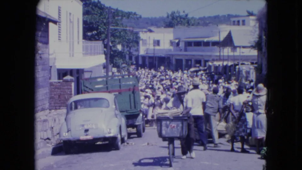 Elevated view of a crowded Kingston street market in 1964, with a Beetle, truck, carts, and pedestrians filling the road.