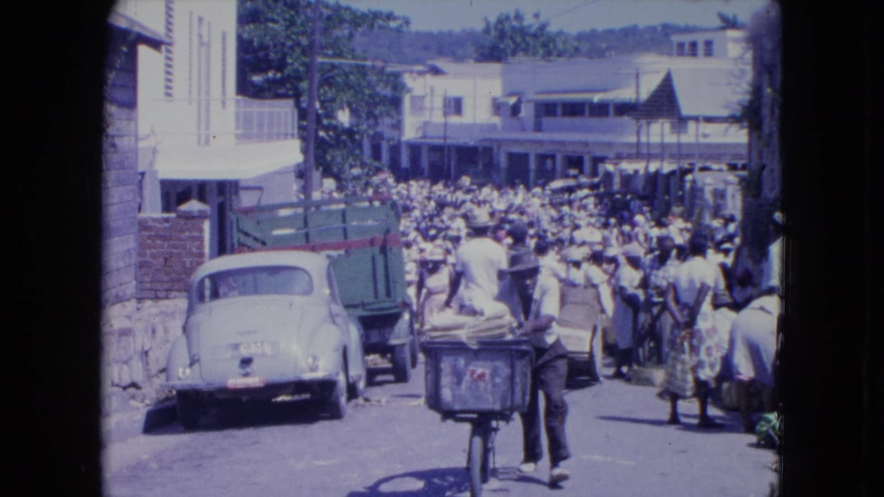 1964 Kingston market street with a bicycle cart in the foreground, parked vehicles, and a dense crowd moving between buildings.