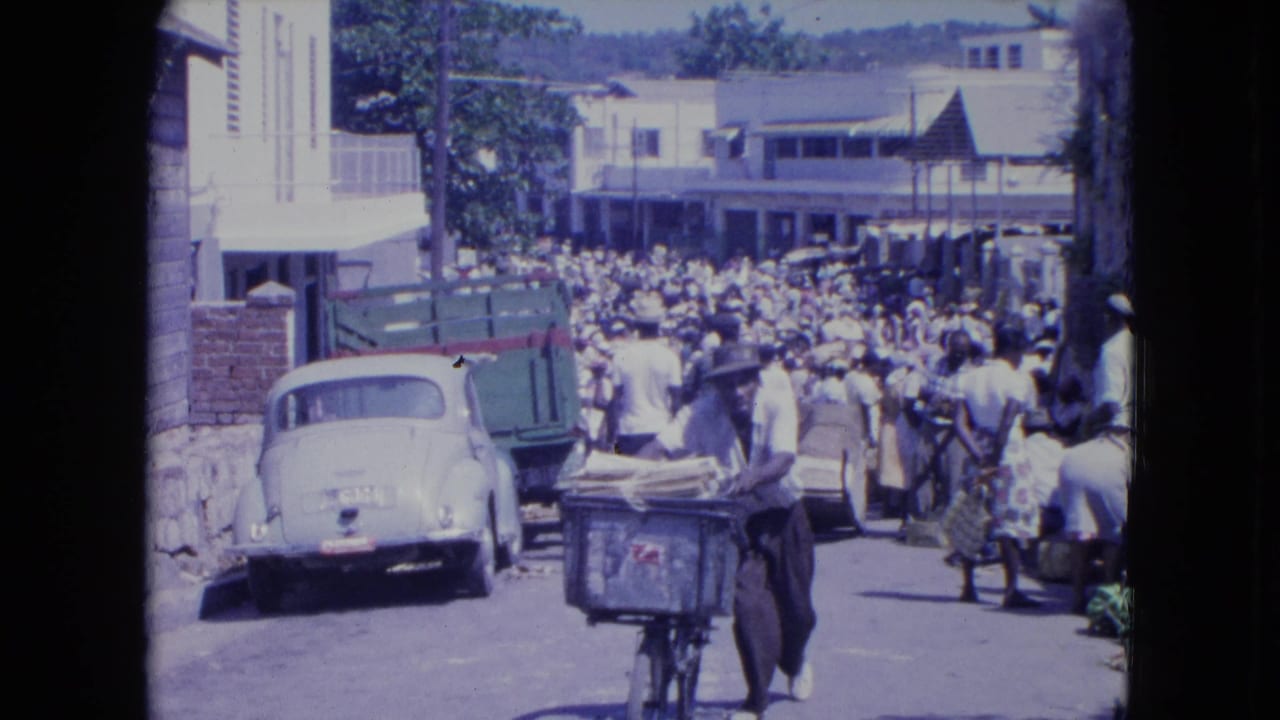 In 1964 Kingston, Jamaica, a man pushes a bicycle stacked with baggage through a crowded street beside a white car and green truck.