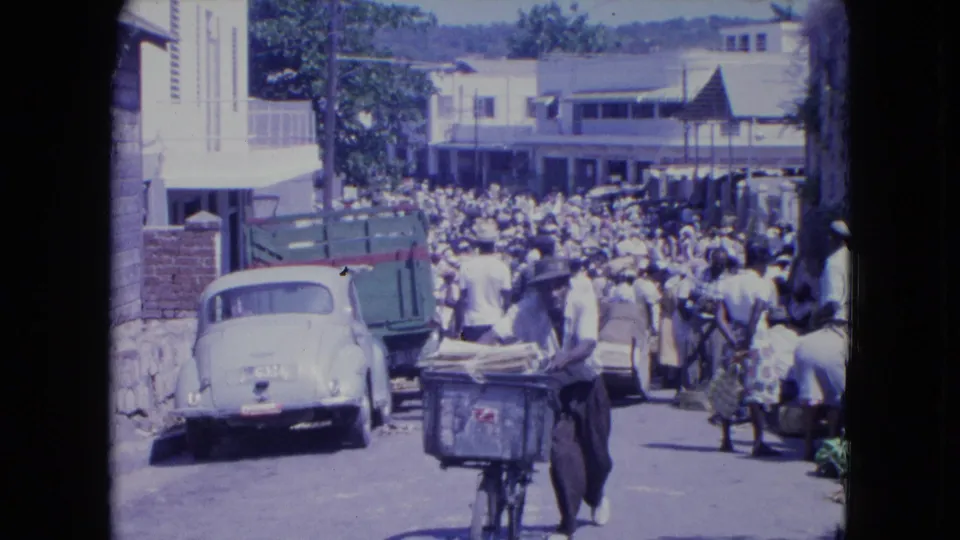 In 1964 Kingston, Jamaica, a man pushes a bicycle stacked with baggage through a crowded street beside a white car and green truck.