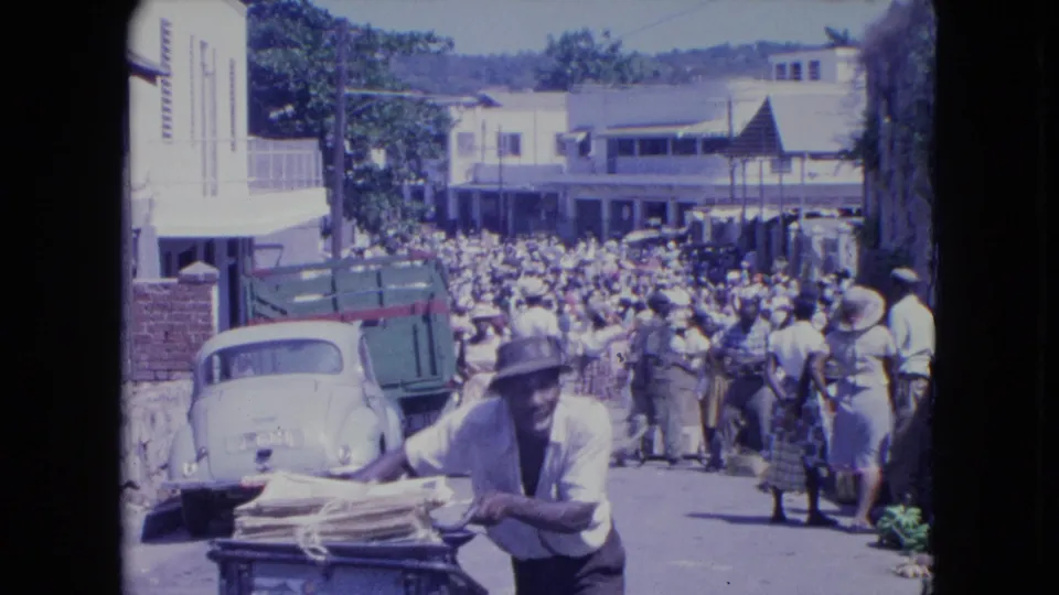 The man in a brimmed hat pushes his loaded bicycle nearer the camera while people and vehicles fill the Kingston street behind him.