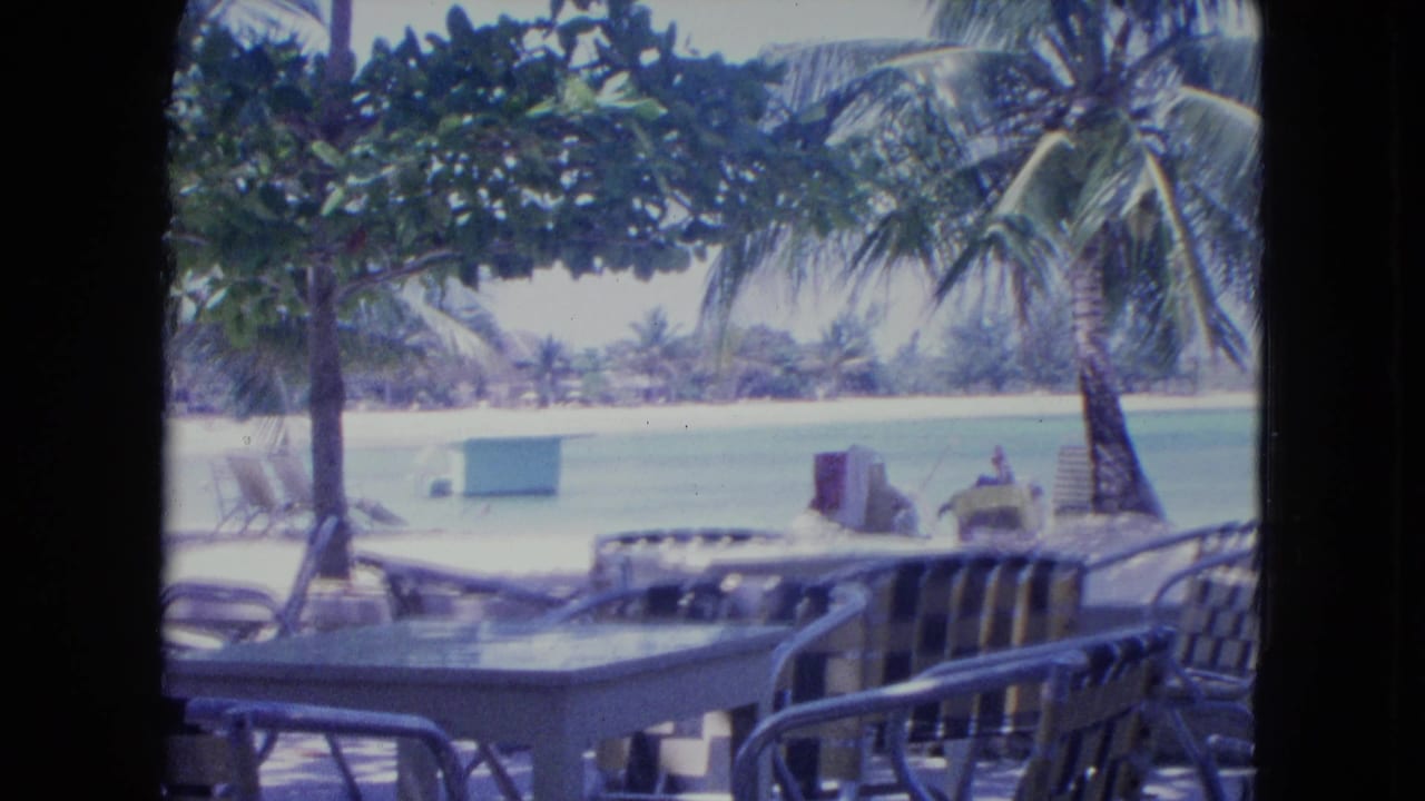 1964 Kingston, Jamaica 8mm frame showing a shaded resort patio with tables, chairs, palms, white sand, and turquoise lagoon water beyond a window-like divider.