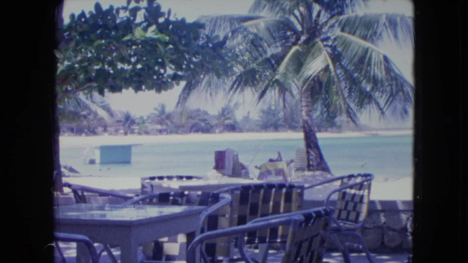 1964 Kingston, Jamaica resort view with shaded metal patio chairs and tables in the foreground, a low wall, palm trees, bright sand, and calm turquoise water in the background.
