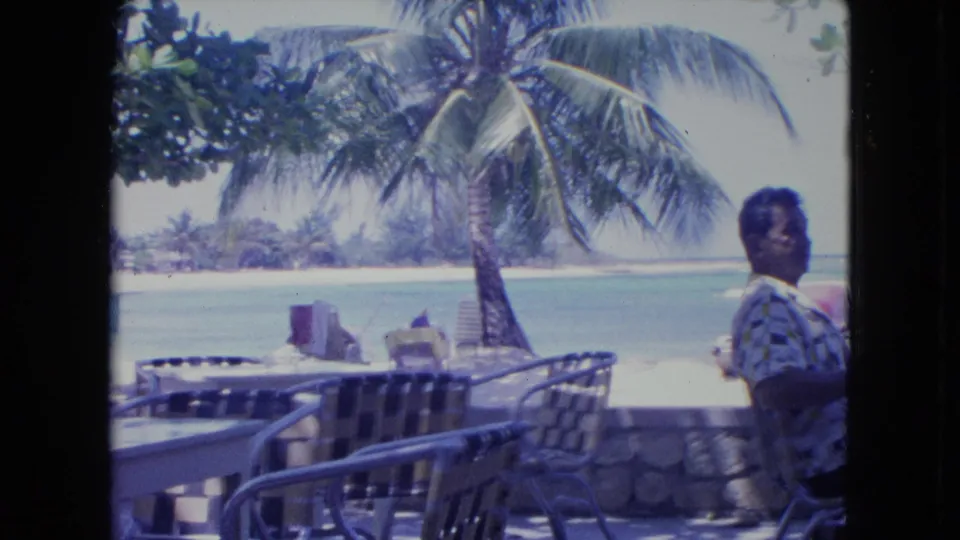 1964 Kingston, Jamaica 8mm frame showing a beachside resort patio, palm trees, lagoon water, and a visible adult figure at the right edge of the frame.