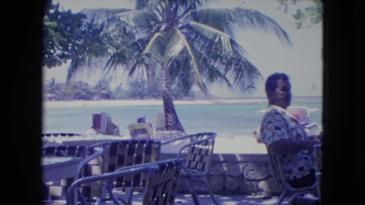 In 1964 Kingston, Jamaica, an older man sits at an outdoor beachside cafe table near woven chairs, a palm tree, and turquoise water.