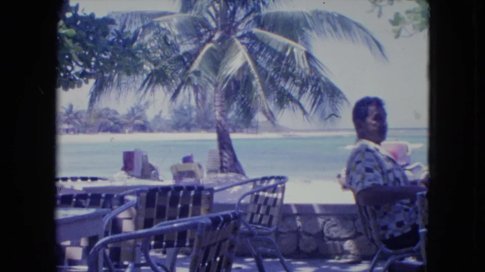 In 1964 Kingston, Jamaica, an older man sits at an outdoor beachside cafe table near woven chairs, a palm tree, and turquoise water.