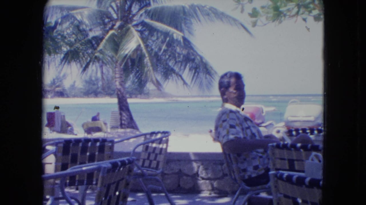 At a Kingston beachside cafe in 1964, the seated man turns toward the camera beside patio furniture, palm fronds, and bright ocean water.