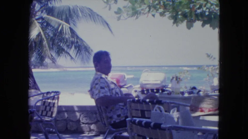 The 1964 Kingston home movie shows a side view of the seated man at a beachside cafe, framed by chairs, a stone wall, palm leaves, and sea.