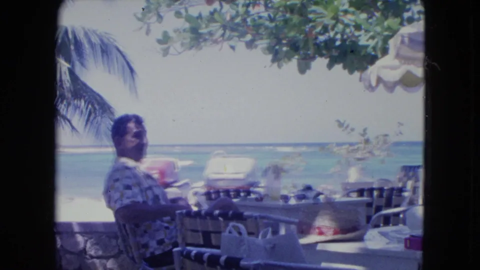 At the end of the 1964 Kingston beachside cafe shot, the older man remains seated near tabletop items, chairs, foliage, and the ocean.