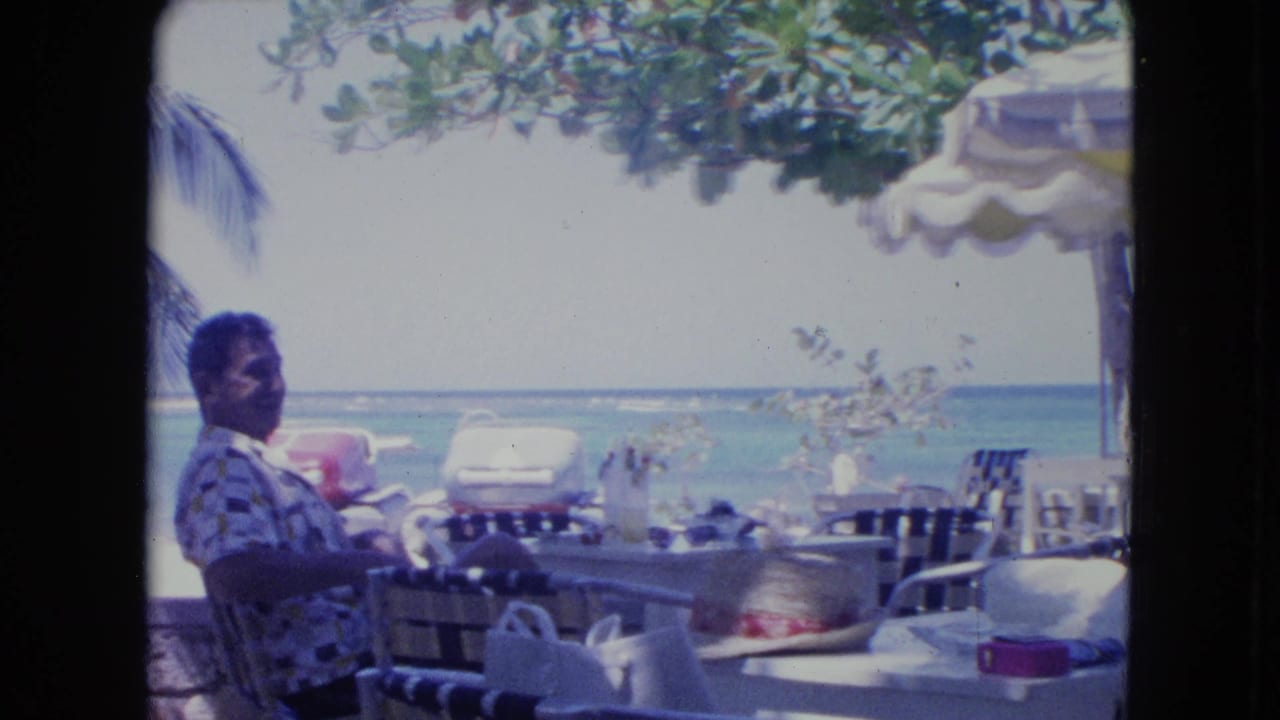 1964 Kingston, Jamaica home-movie frame showing a man at a beachside café table facing the ocean under a striped umbrella.
