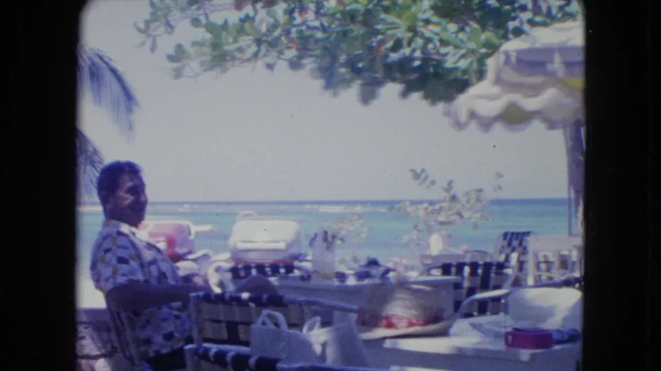 1964 Kingston, Jamaica home-movie frame showing a man at a beachside café table facing the ocean under a striped umbrella.