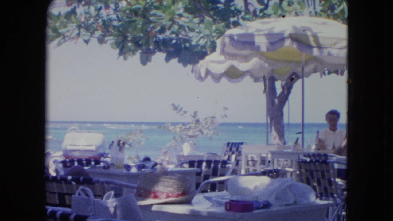 Beachside café tables and chairs under an umbrella, with ocean water and a standing man at right.