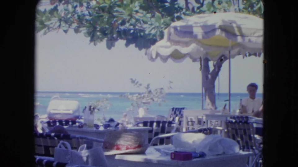 1964 Kingston, Jamaica frame of café seating by the beach, with a striped umbrella, ocean view, and a man standing near the right edge.