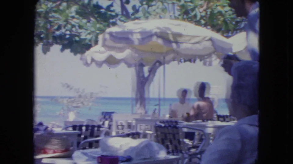 1964 Kingston, Jamaica beach café view with adults seated beneath a striped umbrella and the turquoise water beyond.