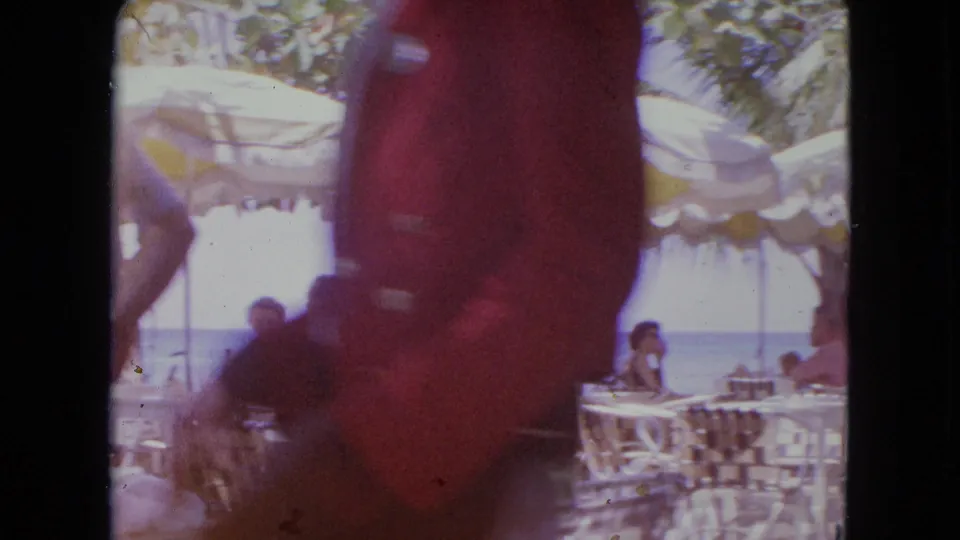 1964 Kingston, Jamaica home-movie frame with a close passerby in red partly obscuring café tables, seated adults, umbrellas, and beach water.