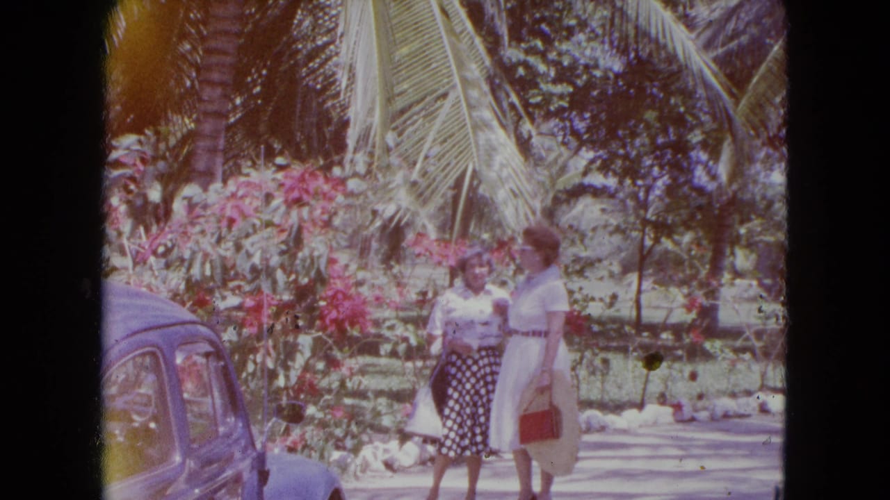 Two adult women in 1964 Kingston, Jamaica, stand beside a blue classic car near flowering shrubs and palm trees.