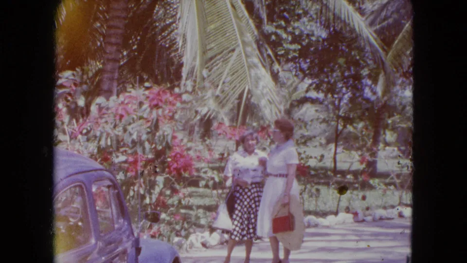 Two adult women in 1964 Kingston, Jamaica, stand beside a blue classic car near flowering shrubs and palm trees.