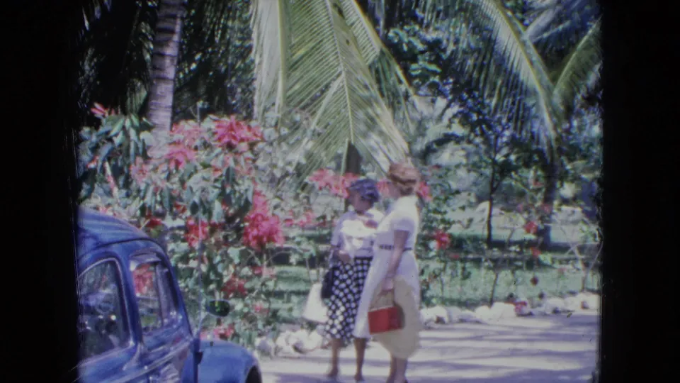 Two adult women walk through a tropical garden in Kingston, Jamaica, with a blue classic car and bright pink flowers nearby.