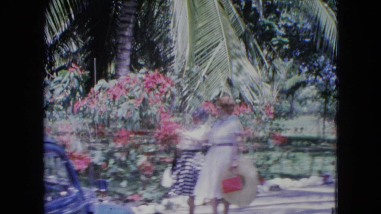 In the 1964 Kingston garden scene, two women move past pink blossoms while carrying a straw hat and red purse.