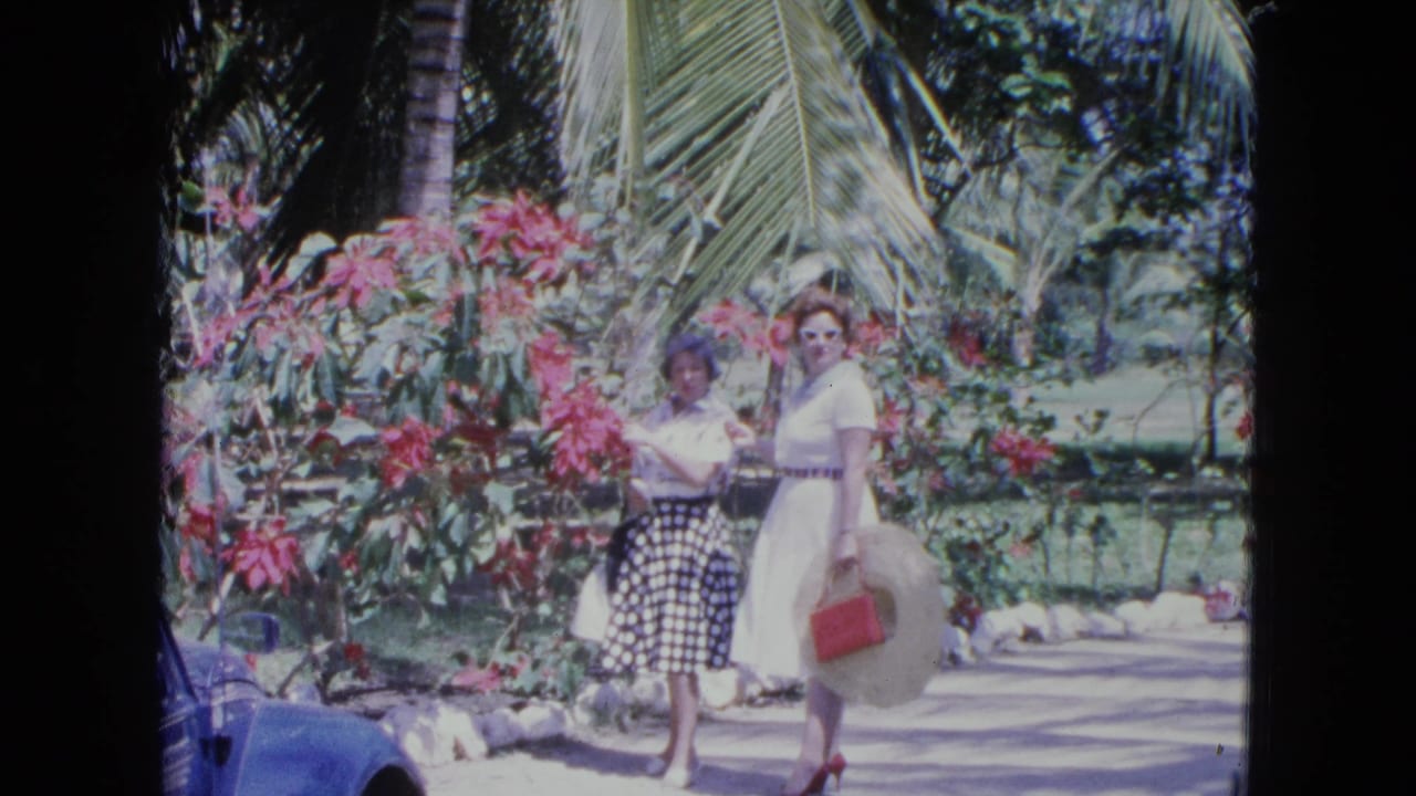 Two women pose for a home-movie camera in a Kingston garden, surrounded by pink blossoms and palm leaves.