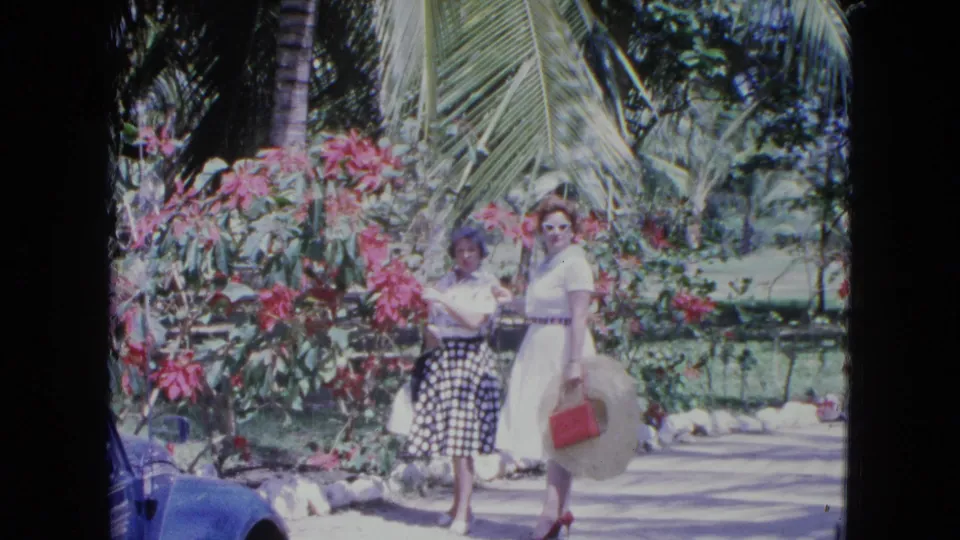 Two women pose for a home-movie camera in a Kingston garden, surrounded by pink blossoms and palm leaves.