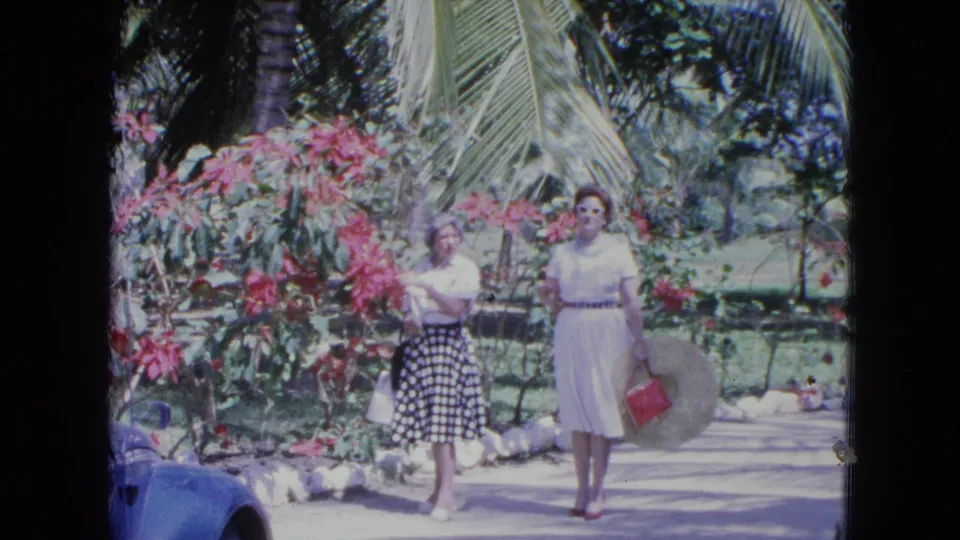 Two women stroll through a Kingston tropical garden, one in a white dress and one in a polka dot skirt.