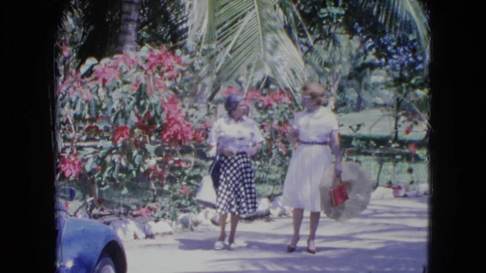 The women continue along a paved path in a Kingston garden, with tropical planting and a partial blue car nearby.