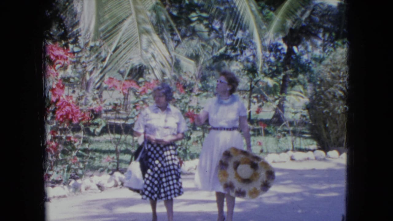 Two adult women stand or walk on a tropical path as one gestures near red flowers.