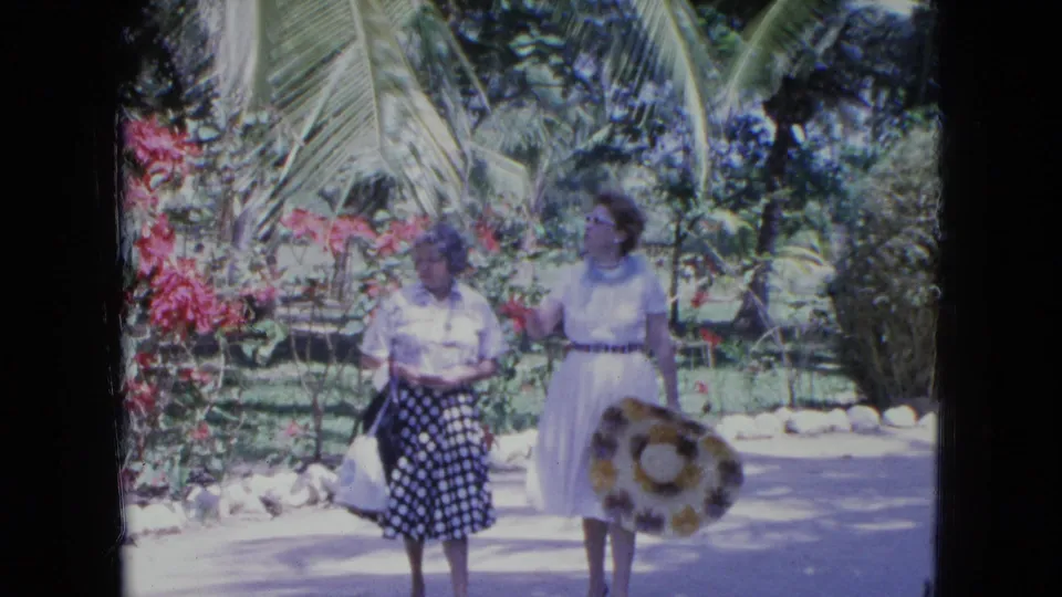 In the Kingston 1964 home-movie scene, two adult women pause or walk slowly, with one gesturing beside palm fronds and red flowering shrubs.
