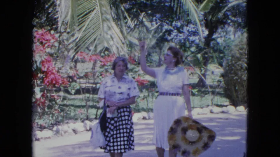 In 1964 Kingston footage, two adult women stand close on a tropical path, one lifting her arm while holding a large straw hat at her side.