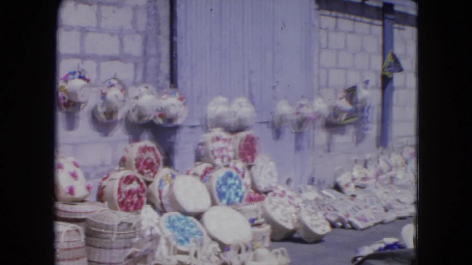 1964 Kingston, Jamaica sidewalk stall showing woven baskets and bright floral hats arranged against a block wall.