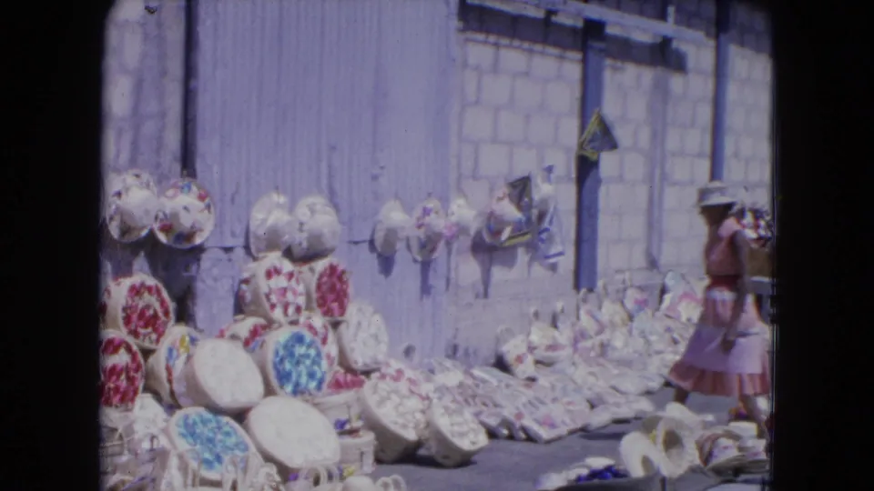 1964 Kingston, Jamaica street-market view with a woman passing a display of handmade baskets and patterned hats.