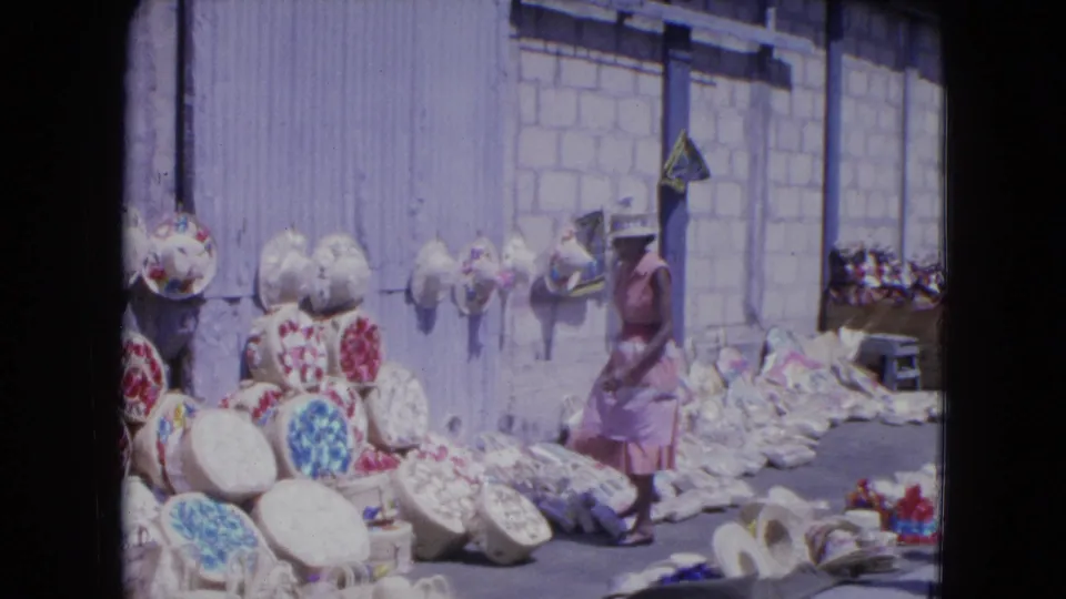 1964 Kingston, Jamaica market scene with a woman near woven baskets, floral hats, and craft goods on the sidewalk.