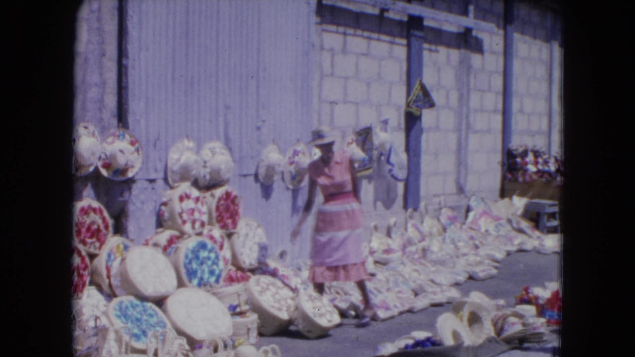 In 1964 Kingston, Jamaica, a woman in a pink dress and hat looks over baskets and woven goods laid out along a street wall.