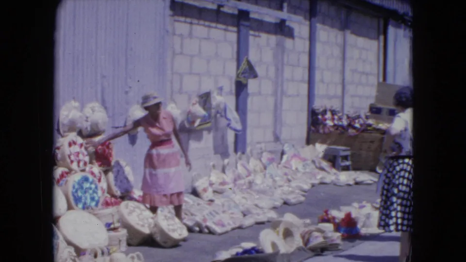 A Kingston street-market view shows a woman in pink gesturing near baskets as another shopper stands beside colorful goods.