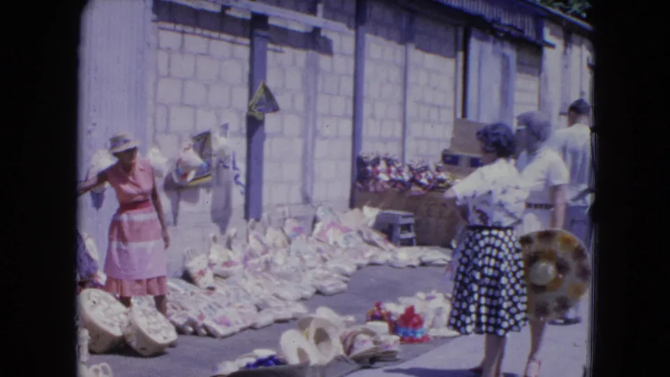 A woman in a polka-dot skirt faces a market display while a woman in pink stands near rows of baskets and hats in Kingston.