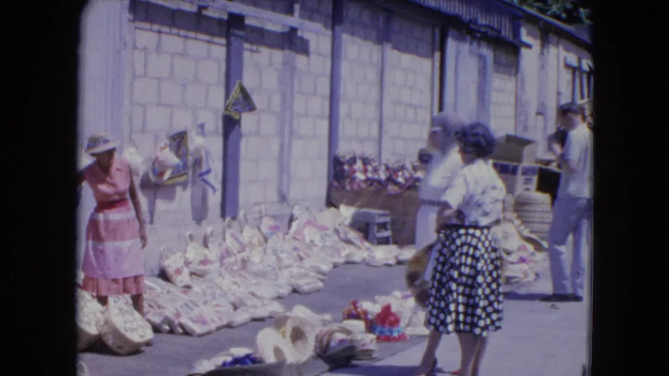 At the Kingston market, a woman in a polka-dot skirt moves past baskets and hats as other adults stand near the street display.